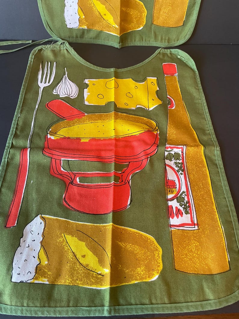 May include: An olive green adult bib featuring a graphic print of a red fondue pot, a piece of yellow cheese, a garlic bulb, a fork, a loaf of bread, and a tall bottle of oil. The design includes bold outlines and vibrant yellow and red accents.