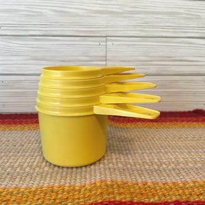 May include: A set of five nested yellow plastic measuring cups stacked vertically. Each cup features a flat handle extending from the side, with the cups decreasing in size from the bottom to the top, creating a uniform, bright yellow kitchen tool set.