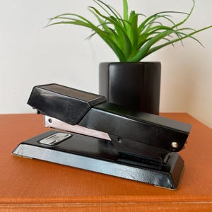 May include: A black stapler with a silver staple guide sits on an orange book. A small, green plant in a black square pot is in the background. The stapler is angled towards the viewer, showcasing its design.