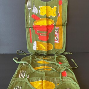 May include: A set of olive green fabric aprons featuring a screen-printed graphic of a fondue pot, a block of Swiss cheese, a clove of garlic, a long fork, a piece of bread, and a bottle of hot sauce. Each apron has matching green fabric ties.