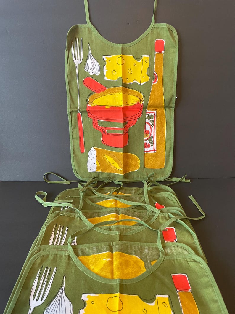 May include: A set of olive green fabric aprons featuring a screen-printed graphic of a fondue pot, a block of Swiss cheese, a clove of garlic, a long fork, a piece of bread, and a bottle of hot sauce. Each apron has matching green fabric ties.