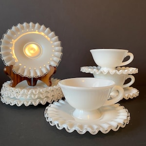 May include: A collection of white milk glass teacups and saucers. The saucers have a ruffled edge with a clear glass trim. The teacups are a simple, classic design. The set includes several saucers and teacups, perfect for a tea party.