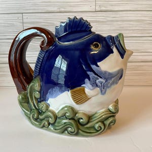 May include: A ceramic teapot shaped like a blue fish with a brown handle. The fish has white and blue accents, a green base with wave details, and a brown handle. The teapot is a decorative kitchen item.