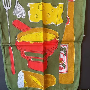 May include: An olive green apron featuring a graphic print of a red fondue pot, a yellow block of Swiss cheese, a long fork, a clove of garlic, a piece of crusty bread, and a tall yellow bottle with a red cap, all outlined in black ink.