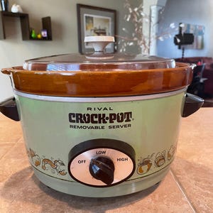 May include: A vintage Rival Crock-Pot Removable Server with a light green exterior, a brown ceramic rim, and a clear glass lid. The front features a black control knob with Off, Low, and High settings, surrounded by small illustrations of food items.