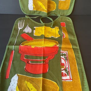 May include: An olive green fabric bib featuring a screen-printed graphic of a red fondue pot, a yellow piece of cheese, a bottle of wine, a fork, a clove of garlic, and a loaf of bread. The bib includes matching green fabric neck ties.