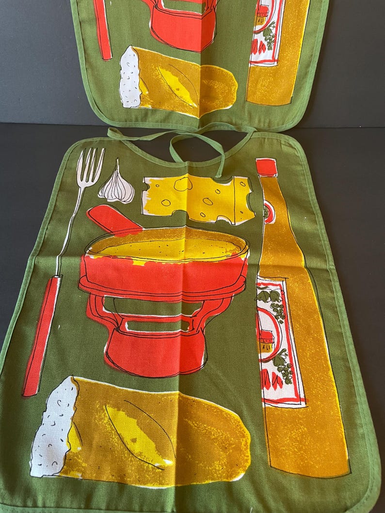 May include: An olive green fabric bib featuring a screen-printed graphic of a red fondue pot, a yellow piece of cheese, a bottle of wine, a fork, a clove of garlic, and a loaf of bread. The bib includes matching green fabric neck ties.