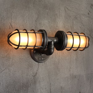 Industrial double cage wall light fixture installed facing upward, displaying frosted glass jelly jar globes casting a soft warm glare-free amber illumination from inside rustic black metal protective pipe cages by MillerLights®.