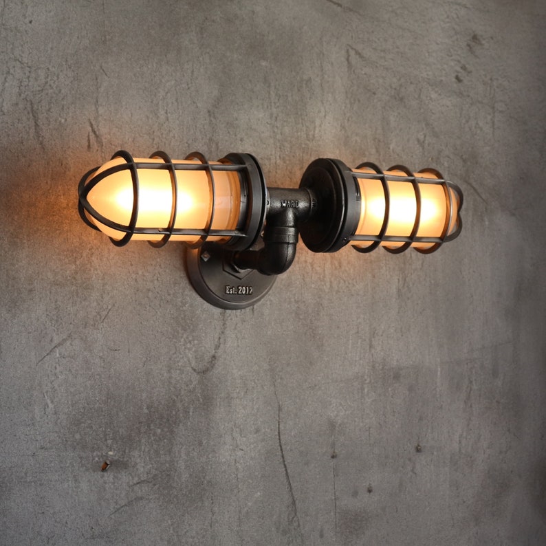Industrial double cage wall light fixture installed facing upward, displaying frosted glass jelly jar globes casting a soft warm glare-free amber illumination from inside rustic black metal protective pipe cages by MillerLights®.