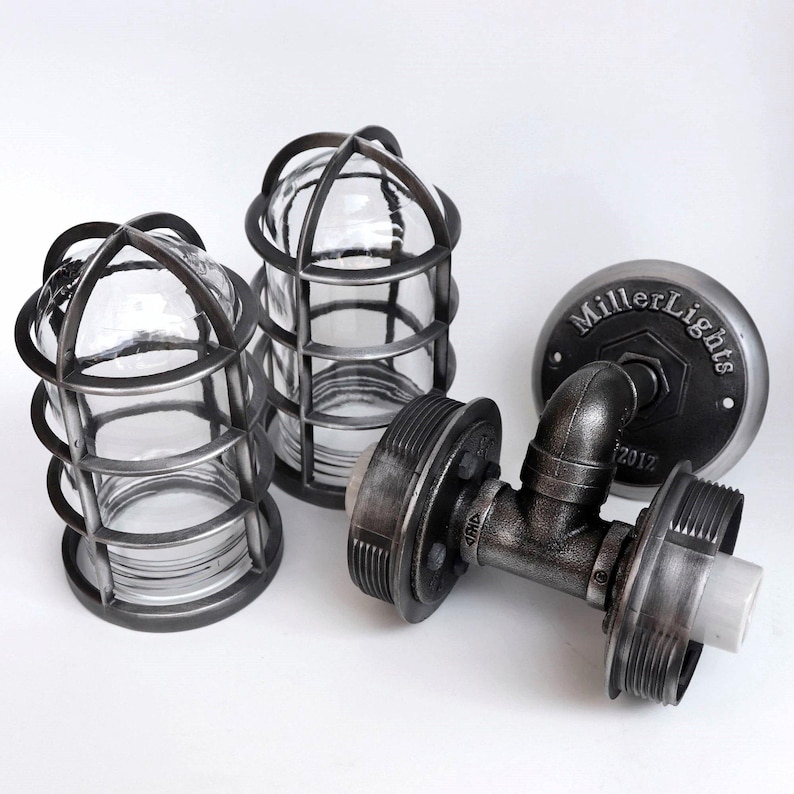 Behind the scenes workshop photo of 'The Joker' double sconce components laid out on a white background, featuring the branded MillerLights® Est. 2012 round iron wall bracket flange, T-pipe body, ceramic bulb sockets, and two detached metal protective cage guards with clear glass shades.