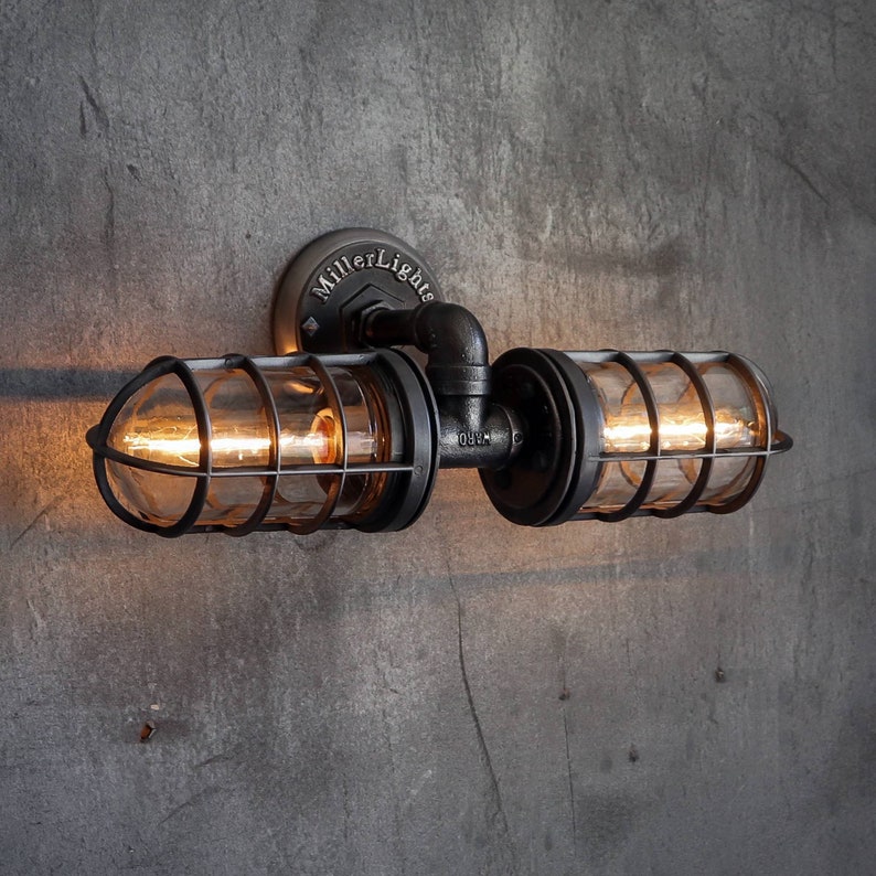 Front view of The Joker model industrial double cage wall sconce lighting fixture by MillerLights®, featuring handcrafted black iron piping, two protective horizontal wire cages over glowing warm Edison bulbs, mounted on a textured concrete wall.