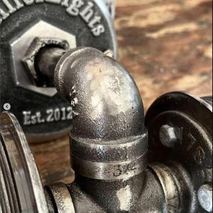 Extreme macro close up of the rugged industrial pipe fittings on a MillerLights® wall sconce, highlighting the dark antiqued steel patina, heavy duty threaded iron joints, etched 3/4 inch size numbers, and a branded round mounting plate showing "Est. 2012".
