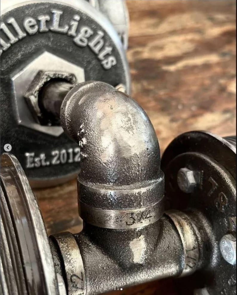 Extreme macro close up of the rugged industrial pipe fittings on a MillerLights® wall sconce, highlighting the dark antiqued steel patina, heavy duty threaded iron joints, etched 3/4 inch size numbers, and a branded round mounting plate showing "Est. 2012".