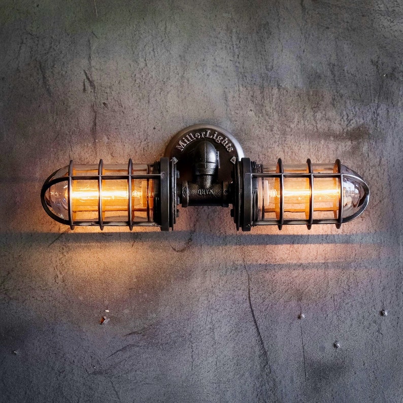 Industrial bathroom vanity light fixture illuminated with a warm amber glow from elongated Edison bulbs, showcasing a black pipe double cage wall sconce with clear glass covers from MillerLights® mounted on a rustic concrete wall.