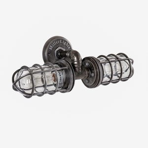Three quarter angle view of The Joker double cage industrial wall sconce by MillerLights®, showcasing unlit clear glass covers, robust black iron pipe plumbing hardware joints, and an antiqued darkened steel finish on a white background.