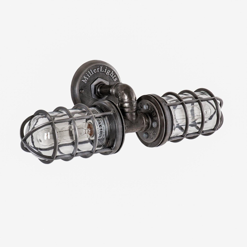 Three quarter angle view of The Joker double cage industrial wall sconce by MillerLights®, showcasing unlit clear glass covers, robust black iron pipe plumbing hardware joints, and an antiqued darkened steel finish on a white background.