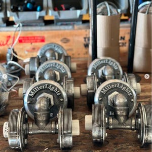 Production line of industrial double cage wall sconce lighting fixtures being handcrafted on a workshop bench, showcasing antiqued steel pipe hardware frames and custom branded MillerLights® round metal backplates.
