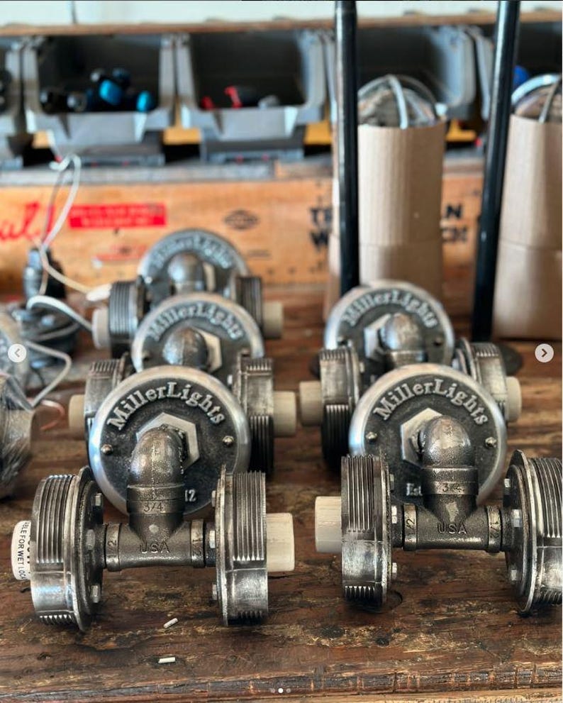 Production line of industrial double cage wall sconce lighting fixtures being handcrafted on a workshop bench, showcasing antiqued steel pipe hardware frames and custom branded MillerLights® round metal backplates.