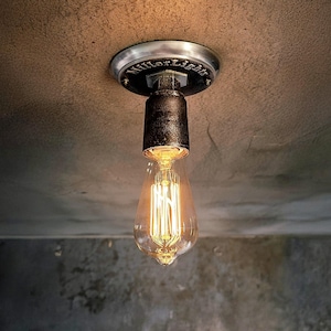 May include: A ceiling-mounted light fixture featuring a dark, textured metal pipe base with the text MillerLights embossed on the circular mounting plate. A clear glass Edison-style bulb with a visible glowing filament hangs from the pipe socket.