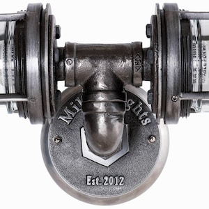 Symmetrical close up view of the black iron T-pipe fitting on a MillerLights® double vanity light, highlighting a branded round mounting base plate showing "Est. 2012" and heavy duty industrial metal craftsmanship on a white background.
