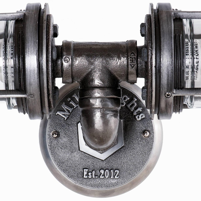 Symmetrical close up view of the black iron T-pipe fitting on a MillerLights® double vanity light, highlighting a branded round mounting base plate showing "Est. 2012" and heavy duty industrial metal craftsmanship on a white background.