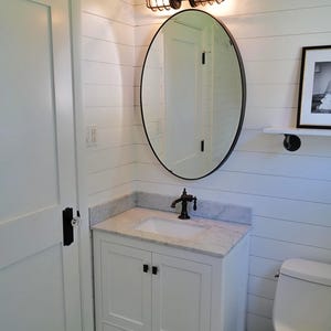 Rustic industrial bathroom vanity light fixture installed above an oval mirror in a modern farmhouse bathroom remodel, casting a warm amber glow over a white single sink vanity cabinet on a shiplap wall by MillerLights®.
