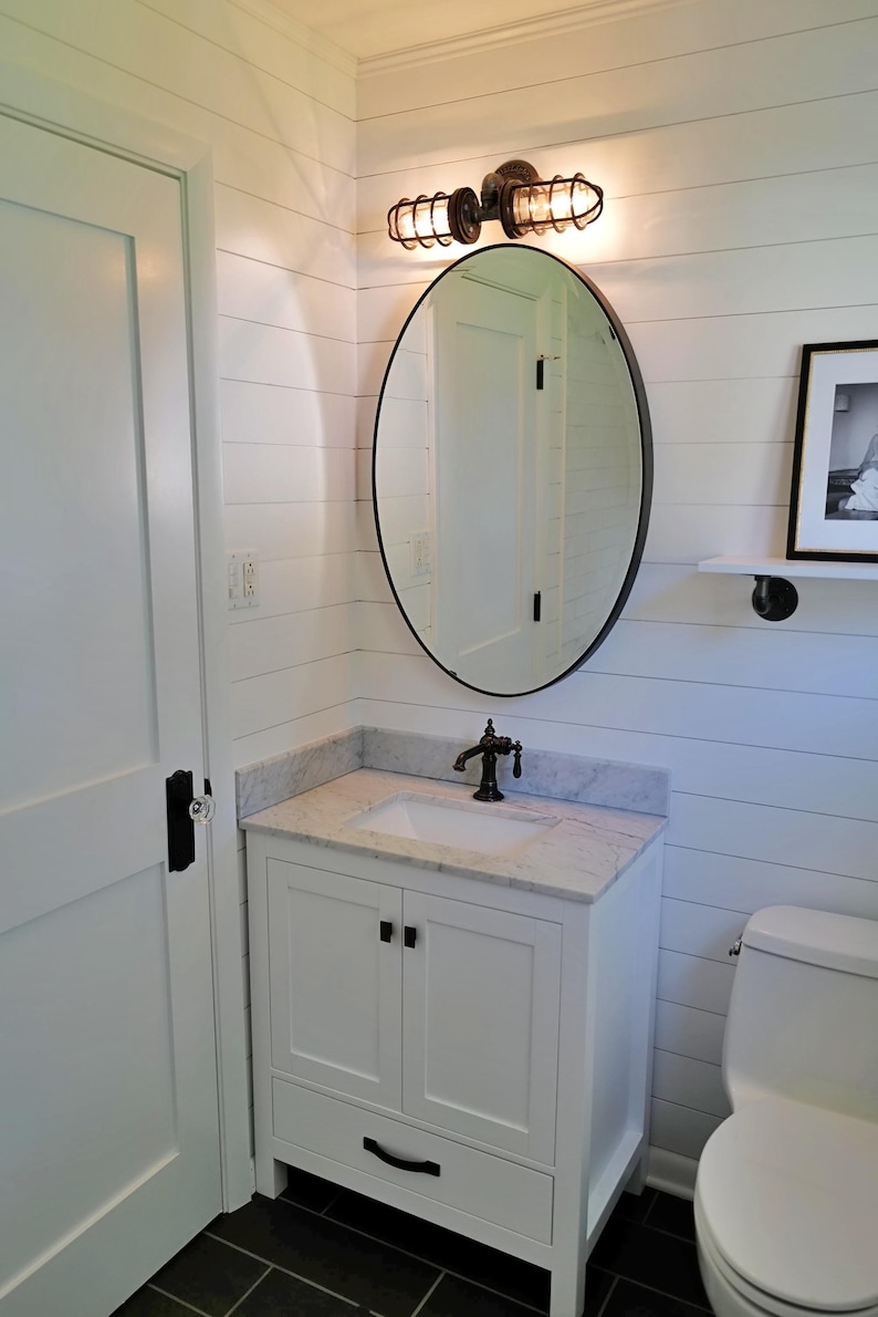 Rustic industrial bathroom vanity light fixture installed above an oval mirror in a modern farmhouse bathroom remodel, casting a warm amber glow over a white single sink vanity cabinet on a shiplap wall by MillerLights®.
