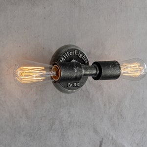 May include: A black metal wall sconce with two Edison bulbs. The sconce has a round base with the text "Miller Lights Est. 2012".