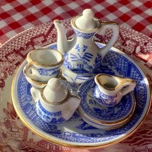 Puede incluir: Juego de té de porcelana en miniatura con diseño Willow en azul y blanco. El juego incluye una tetera, una jarrita para la crema, un azucarero y dos tazas, todo sobre un plato a juego con borde dorado. El juego se exhibe sobre un plato más grande con un patrón rojo y blanco.