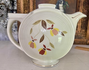 May include: Cream-colored ceramic teapot with a lid, handle, and spout. The teapot features a floral design in shades of brown, yellow, and red. The base and lid are trimmed with gold. The teapot has a circular shape with ridged detailing.