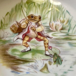 May include: A whimsical illustration on a white surface depicts a frog in a coat and trousers fishing in a pond. The frog stands on a lily pad, holding a fishing rod, with a basket at its side. The scene includes water lilies and a fish.
