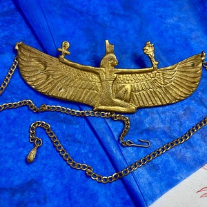 Könnte beinhalten: Eine goldfarbene Halskette mit ägyptischem Anhänger. Der Anhänger zeigt eine geflügelte Figur mit ausgestreckten Armen, die Symbole hält. Die Halskette hat eine Kette mit dekorativem Anhänger und Verschluss. Der Hintergrund ist blau.