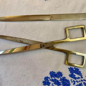 May include: A vintage set of stationery tools, including a letter opener and scissors. The letter opener is a long, rectangular shape with a pointed end. The scissors have gold-colored handles with square finger holes and silver blades.