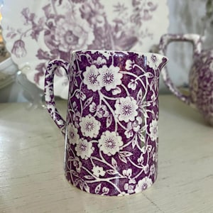 Puede incluir: Una jarrita de cerámica para crema con un patrón floral de transferencia en color morado oscuro y blanco. La jarra tiene un asa curva y un pequeño pico vertedor, con un diseño botánico continuo de flores y hojas que cubre toda la superficie exterior.