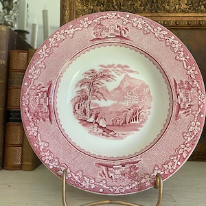 May include: A pink and white ceramic plate with a detailed floral border and a central image of a pink and green landscape with a small cottage. The plate is on a gold metal stand.
