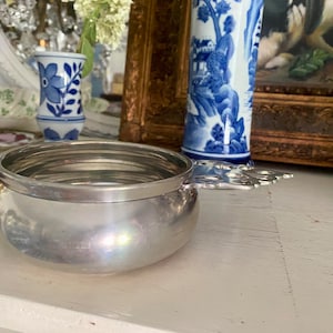 May include: A small, silver-colored bowl with a decorative handle. The bowl has a rounded shape and a smooth, reflective surface. The handle features a floral design. The bowl is likely used for serving or display.