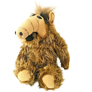 May include: A plush toy character with long, shaggy brown fur, a tan snout, and dark brown hair on top. The creature features a prominent nose with two black nostrils, small black eyes, and a wide mouth with visible black fabric detailing.