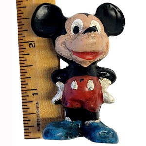 May include: A small, painted figurine of a cartoon mouse with large black ears, a tan face, red shorts with white buttons, and blue shoes. A wooden ruler is positioned vertically next to the figure, showing a height of approximately 2.5 inches.
