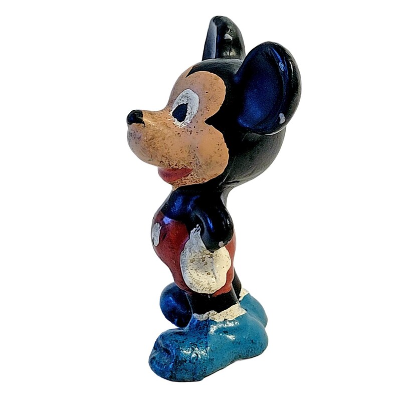 May include: A small, vintage-style figurine of a cartoon mouse character. The figure has a black head and body, a tan face, red shorts with white buttons, and large blue shoes. The paint shows signs of wear and texture throughout the surface.