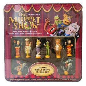 May include: A square metal tin containing The Muppet Show Deluxe Collector's Chess Set. The packaging features red curtains and Muppet characters. Inside, molded plastic holds various Muppet-themed chess pieces, including figures of Kermit, Miss Piggy, and Fozzie Bear, with text detailing the set and age recommendation.