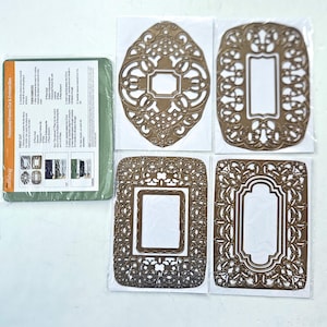 May include: A set of four bronze-colored metal cutting and embossing dies featuring intricate filigree frame designs, accompanied by a green rectangular rubber embossing mat with printed instructional text for crafting applications.
