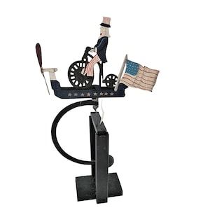 May include: A patriotic folk art sculpture featuring a figure resembling Uncle Sam riding a bicycle. The sculpture includes a propeller, an American flag, and is painted in red, white, blue, and black. The base is black.