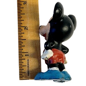 May include: A small, painted figurine of a cartoon mouse character with black ears, a tan face, red shorts, and blue shoes. The figurine is positioned next to a wooden ruler, showing a height of approximately 3 inches from base to ear top.