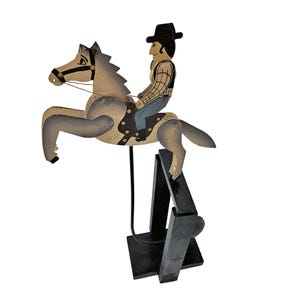 May include: A wooden toy depicting a cowboy riding a horse. The horse is painted in shades of gray and beige, with a black bridle. The cowboy wears a black hat, plaid shirt, and blue pants. The toy is mounted on a black stand.