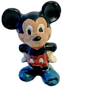 May include: A vintage-style molded figurine of a cartoon mouse character with large round ears. The figure features a black head and torso, a tan face, red shorts with white buttons, white gloves, and large blue shoes, showing signs of paint wear.
