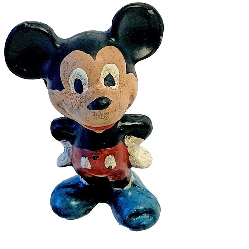 May include: A vintage-style molded figurine of a cartoon mouse character with large round ears. The figure features a black head and torso, a tan face, red shorts with white buttons, white gloves, and large blue shoes, showing signs of paint wear.