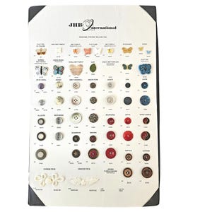 May include: A JHB International seasonal preview display card featuring a collection of assorted buttons in various shapes, sizes, and colors, including butterfly, leaf, and circular designs, with printed labels and style numbers for each item.