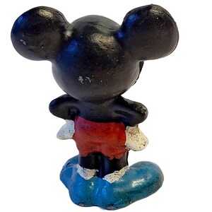 May include: A small, painted figurine of a cartoon mouse character with large black ears, a black head and torso, red shorts, white gloves, and blue shoes. The figure is standing on a blue base with visible paint wear on the ears and gloves.