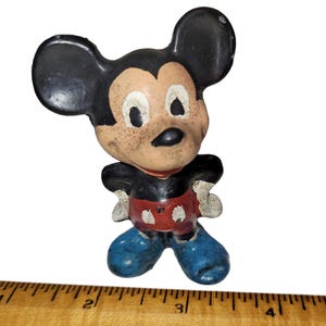 May include: A vintage Mickey Mouse figurine with a black head and ears, a tan face, a black torso, red shorts with white buttons, and blue shoes. The paint shows significant wear and aging. A wooden ruler is visible at the base for scale.