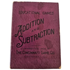 May include: A rectangular purple box for a card game titled EDUCATIONAL GAMES ADDITION AND SUBTRACTION. The text includes COPYRIGHTED, 1902, BY THE CINCINNATI GAME CO. CINCINNATI, O. The box shows signs of wear and discoloration on the surface.